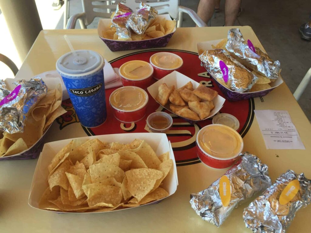 A table filled with fast food items, including trays of tortilla chips, foil-wrapped burritos, cups of cheese dip, and a blue drink cup, with a Taco Cabana review receipt visible on the side.