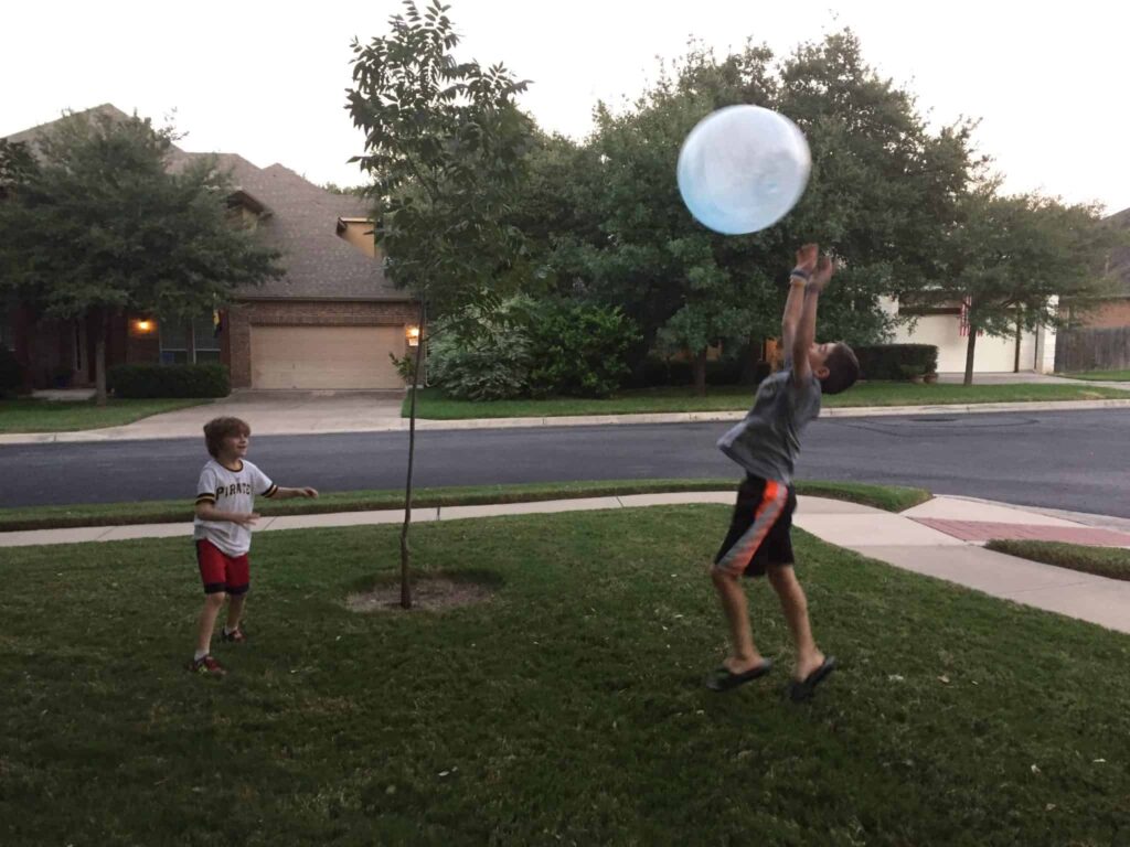Two boys playing with a large blue balloon on a lawn in front of houses. One boy is jumping to catch the Super Wubble Ball while the other stands nearby watching. Perfect for an impromptu play session, this scene could easily be part of a fun Super Wubble Ball review.