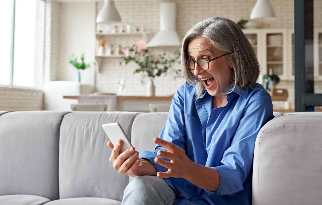 An older woman is sitting on a couch and using a cell phone for buying on eBay.