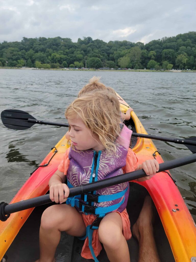 Two children wearing life jackets sit in a tandem kayak on a lake, with a forested shoreline in the background, showcasing one of the many things to do in nature.