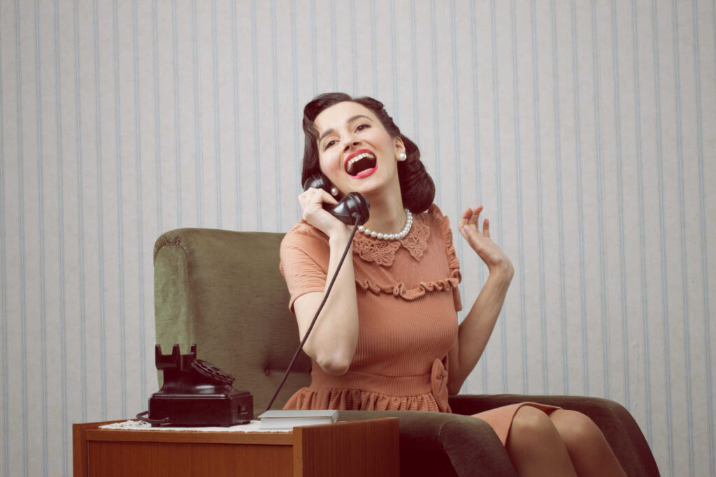 A woman in vintage attire laughs while talking on a rotary phone, seated in an armchair, blending traditions with modern lives effortlessly.