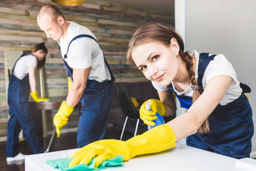 Three people wearing uniforms and yellow gloves are cleaning a room using professional cleaning hacks. One person in the foreground is wiping a surface, while the others are vacuuming and wiping a table in the background.