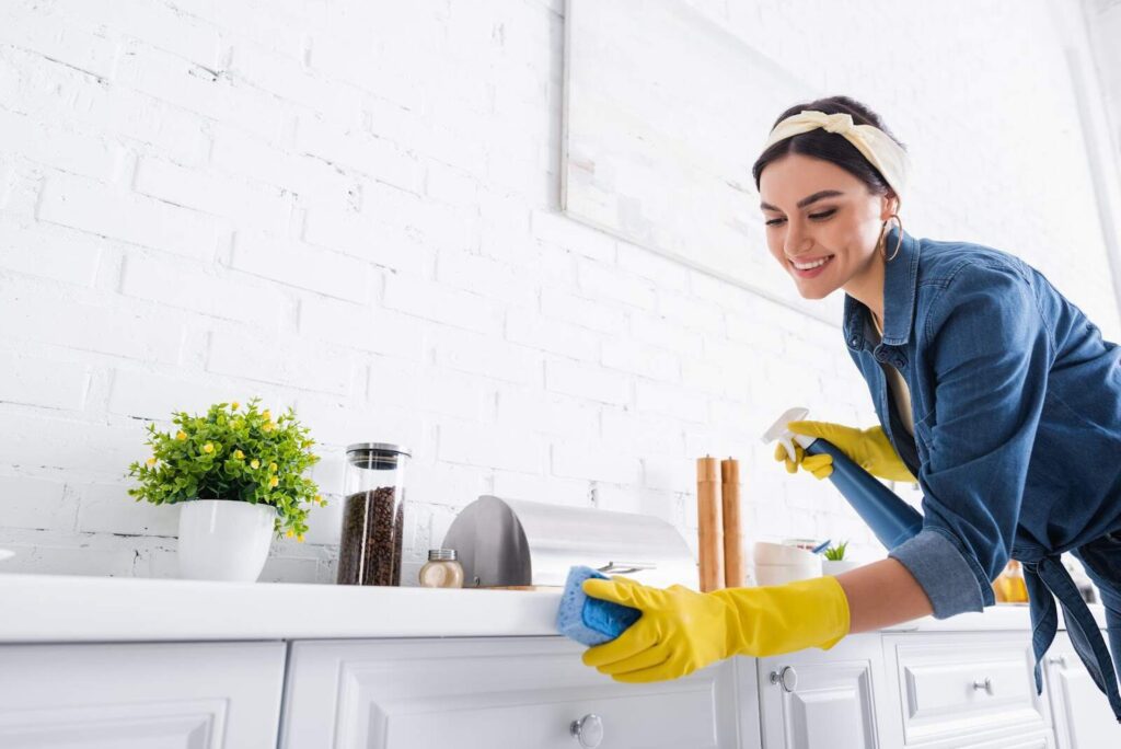 A woman wearing gloves and a headband cleans a kitchen counter with a rag, smiling as she eliminates germs in her bright, white-tiled room.