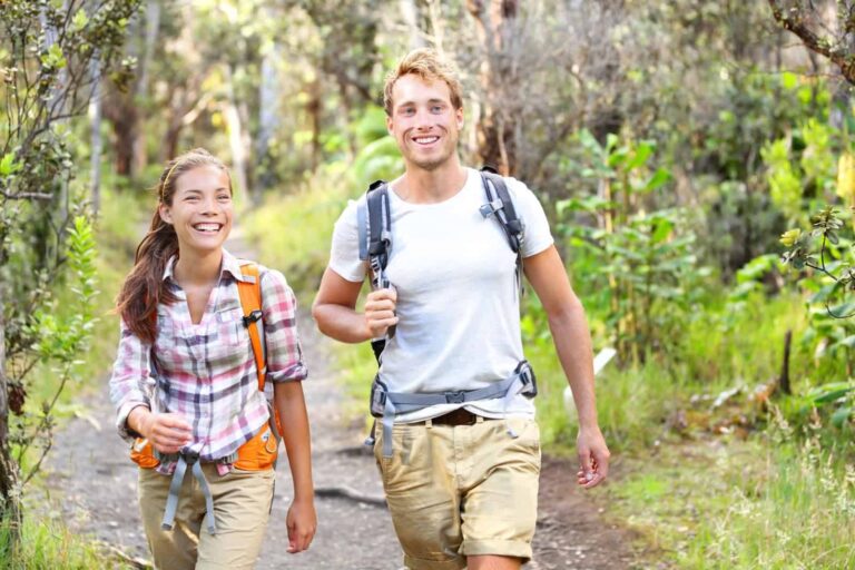 Two happy young hikers with backpacks walking on a forest trail, smiling and looking towards the camera, enjoying things to do in nature.