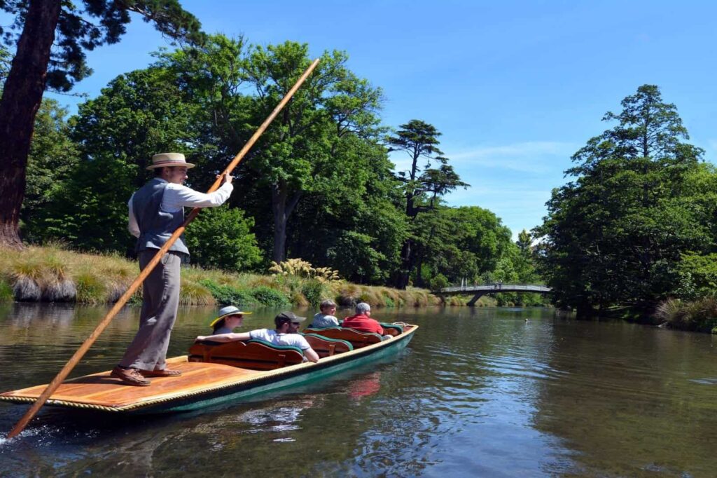 A person standing on a punt navigates a group of people down a calm river surrounded by lush greenery and trees on a sunny day, offering one of the best things to do in nature.
