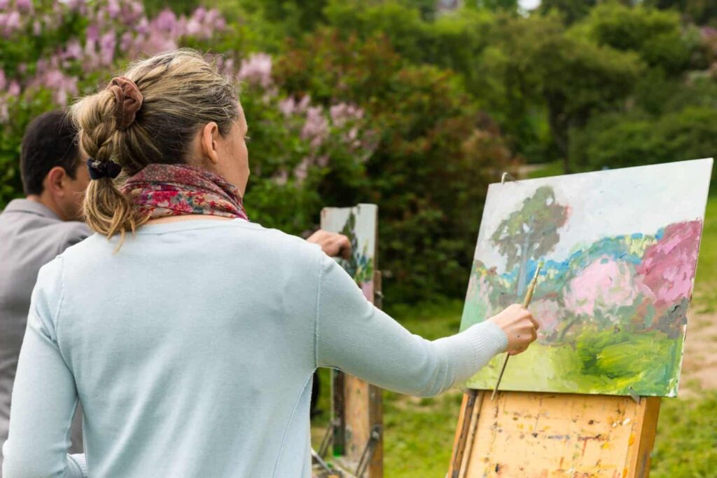 Two people are painting landscape scenes outdoors on easels. The focus is on a woman applying brush strokes to a canvas depicting a natural setting, one of the many enriching things to do in nature.