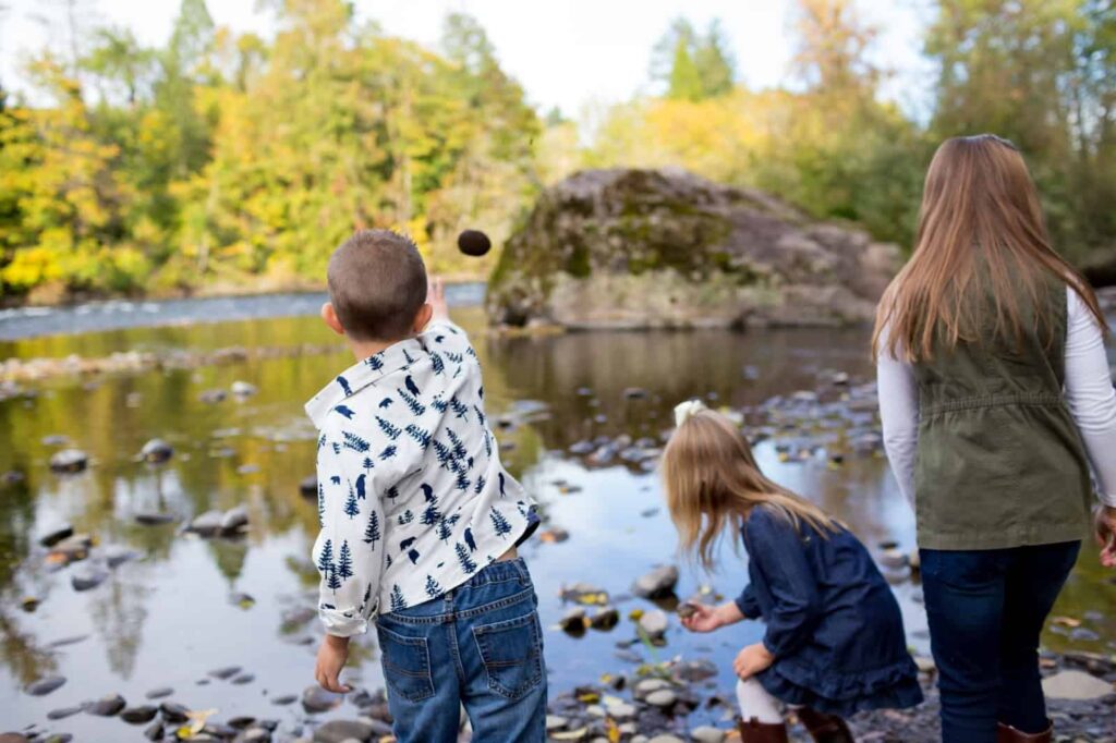 Three children are enjoying things to do in nature by a riverside. One child is throwing a rock into the water, while another is crouching and the third is standing, all facing the river. Trees and rocks surround the scene.