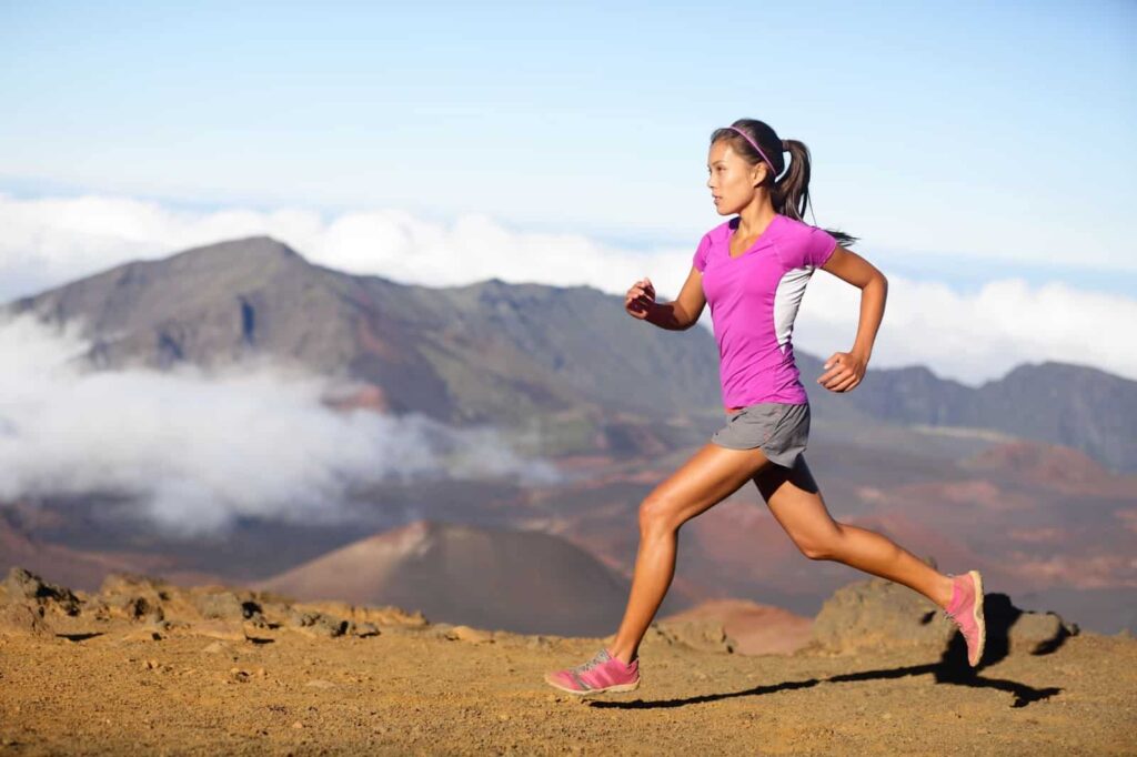 A woman wearing a pink shirt and gray shorts runs on a rocky path on a mountain, showcasing one of the exhilarating things to do in nature, with a scenic landscape of mountains and clouds in the background.