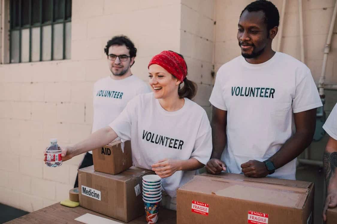 Three volunteers wearing white "Volunteer" T-shirts stand behind a table with cardboard boxes labeled "Medicine" and "Fragile." One woman hands a bottled water across the table, reminding us of the essential things not to leave behind when you die.