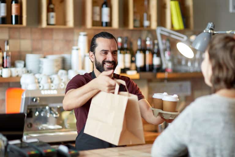 A barista hands a paper bag and tray with two coffee cups to a customer at a cafe counter, mindful that many Americans are tired of tipping.