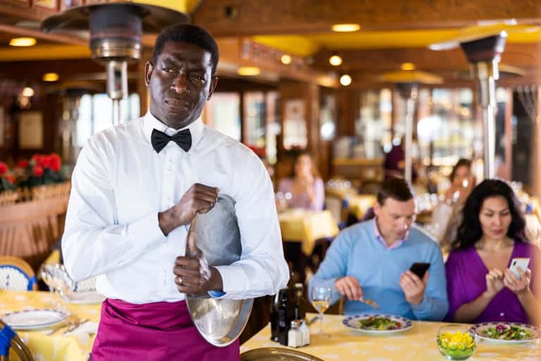 A waiter wearing a white shirt and bow tie holds a silver tray at a restaurant. In the background, a man and woman are seated at a table looking at their phones, perhaps reflecting how Americans are tired of tipping in an increasingly digital age.