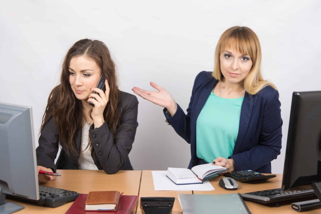 Two women working at desks; one is on the phone and using a pen, while the other gestures with one hand and holds a notebook in the other. Despite being surrounded by digital technology like computers, they embrace traditional office items to combat the bad effects of digital technology.