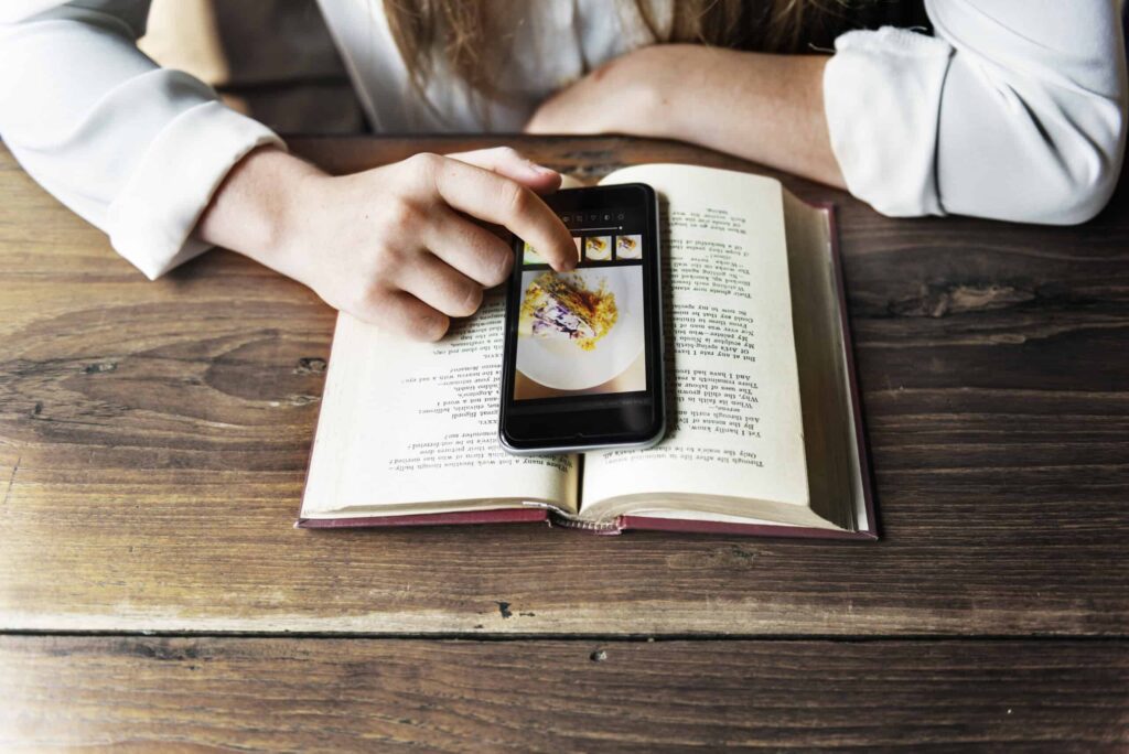 A person using a smartphone to edit a photo of food placed over an open book on a wooden table, showcasing how everyday things are replaced by technology in modern life.