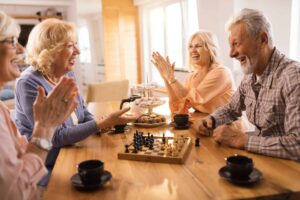 Four elderly individuals are seated around a wooden table, smiling and clapping, with a chessboard and cups of coffee in front of them, reminiscing about things that are obsolete.