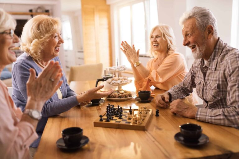 Four elderly individuals are seated around a wooden table, smiling and clapping, with a chessboard and cups of coffee in front of them, reminiscing about things that are obsolete.
