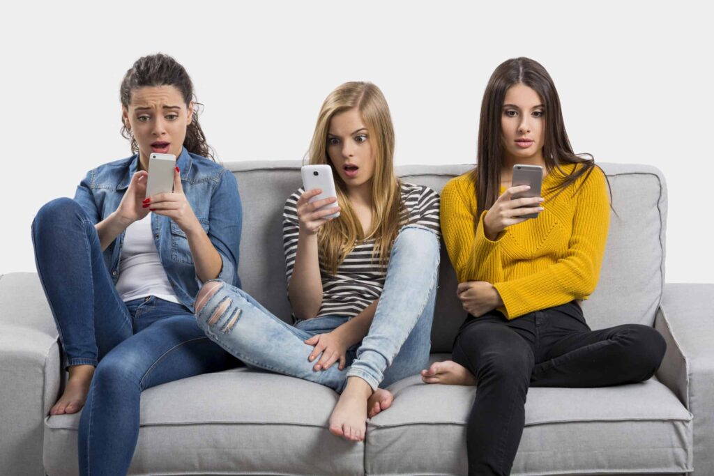 Three young women sitting on a couch, each holding a smartphone with surprised and focused expressions, marvel at how quickly things are replaced by technology.