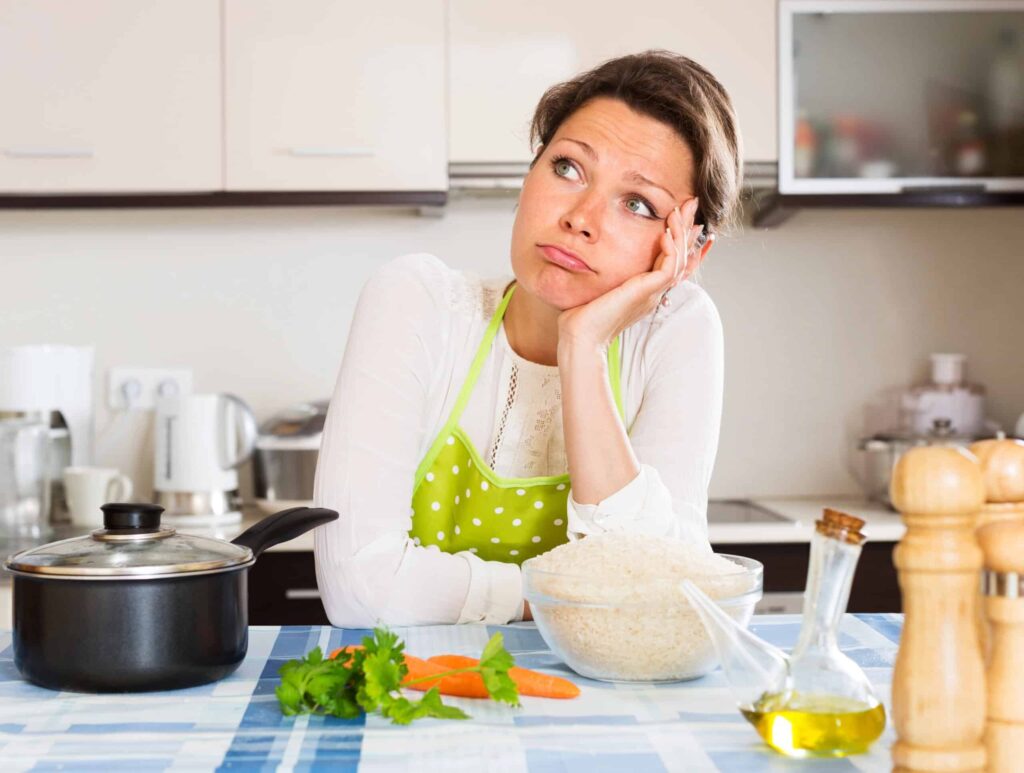 A woman in a green apron, typical of a lower middle income class household, leans on a kitchen counter, looking thoughtful. In front of her are a pot, a bowl of rice, vegetables, and bottles of oil and vinegar.
