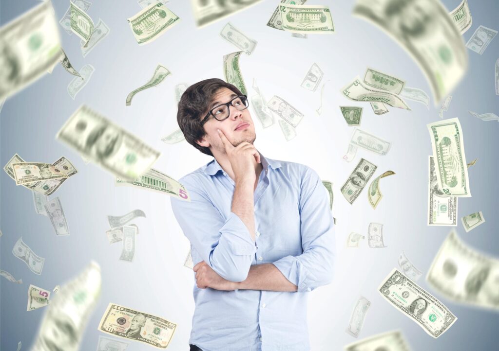 A man in a blue shirt and glasses is looking up thoughtfully, surrounded by falling U.S. dollar bills against a gray background, representing upper class earnings.