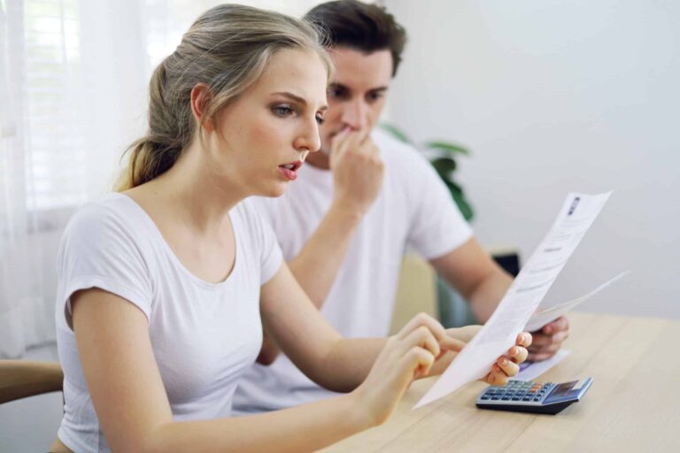 A woman reads a document on how to lower utility bills while a man looks concerned in the background. They are seated at a table with a calculator.