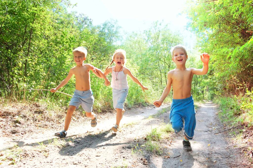 Three children are running happily on a dirt path in a wooded area, reminiscent of childhood in the 1960s. Two boys are shirtless and wear shorts, while the girl in the middle wears a sleeveless top and skirt.