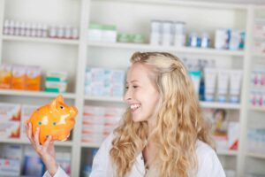 A woman with long blonde hair and a white coat smiles while holding an orange piggy bank in a pharmacy, highlighting how you can save money on prescriptions.