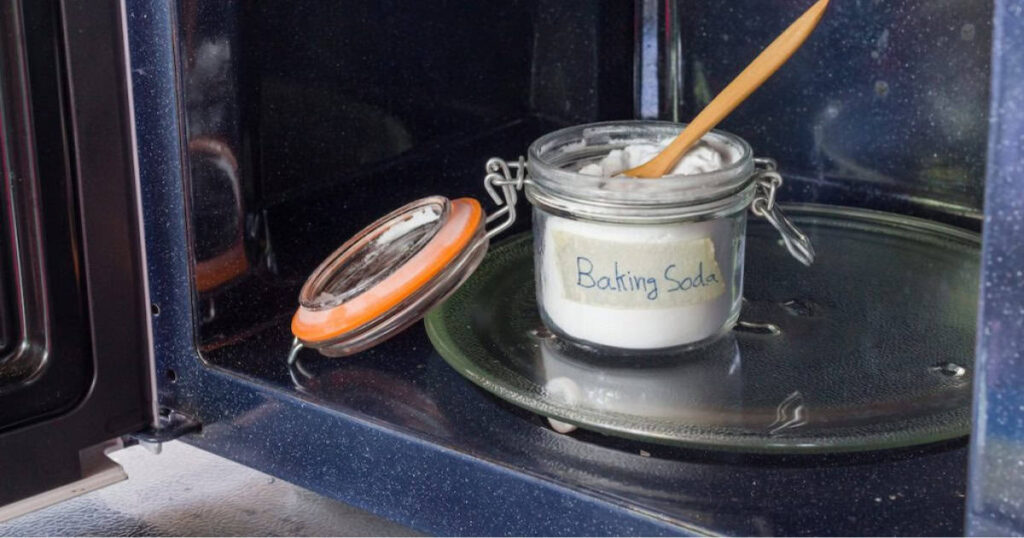 A jar labeled "Baking Soda" with a wooden spoon inside sits on a microwave's rotating glass plate.