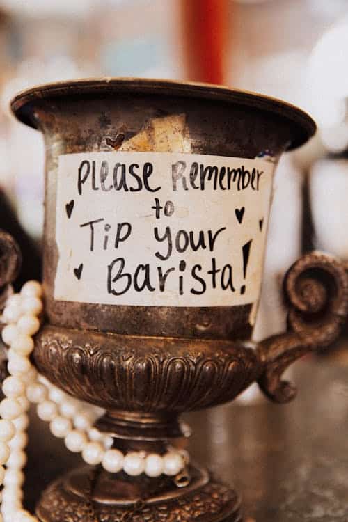 Ornate metal cup with a handwritten sign attached that reads "Please remember to tip your barista!" surrounded by pearl beads, ensuring you don't succumb to tipping fatigue.