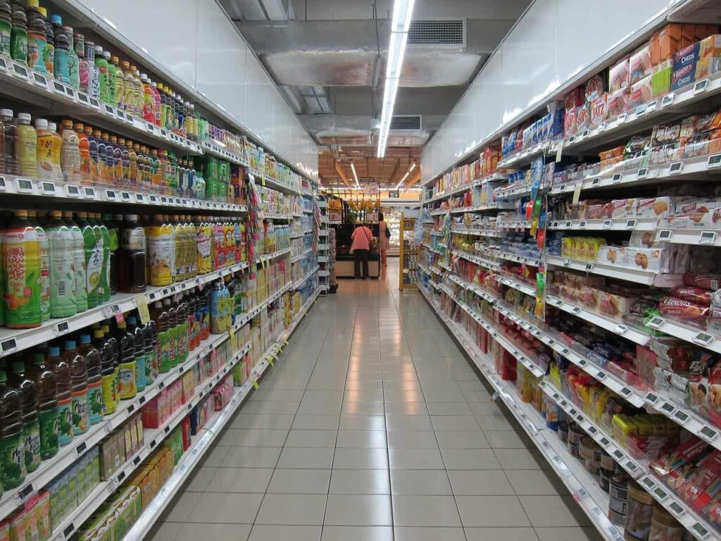 A person with a shopping cart walks down an aisle lined with various packaged food and drink products in a well-lit grocery store, deep in thought about how Americans are tired of tipping everywhere they go.