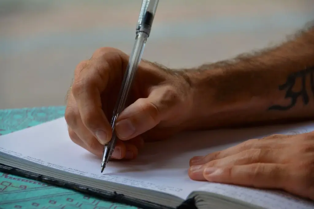 A person with a tattooed arm writes with a pen in a notebook on a table covered with a patterned cloth.