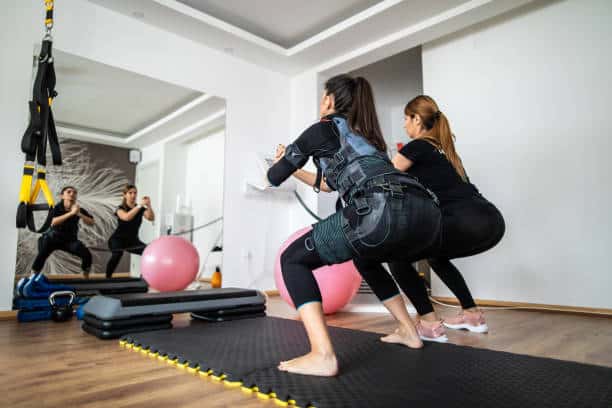 Two women are performing squats on exercise mats in a gym, using suspension trainers. They are facing a mirror, and various fitness equipment is visible in the background.