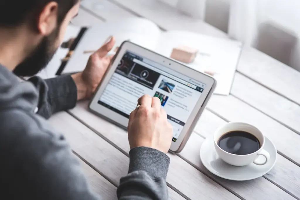 A person is using a tablet with a stylus next to an open book and a cup of coffee on a wooden table, illustrating how things replaced by technology still coexist in daily life.