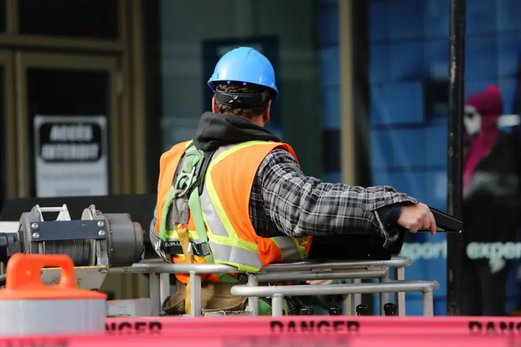 A working-class construction worker in a blue hard hat and orange safety vest operates machinery at a site marked with "Danger" tape.