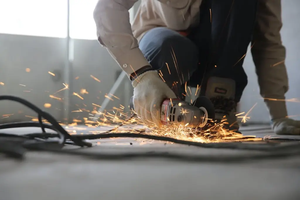 A working-class individual wearing gloves uses an angle grinder, generating sparks in a bustling workshop setting.