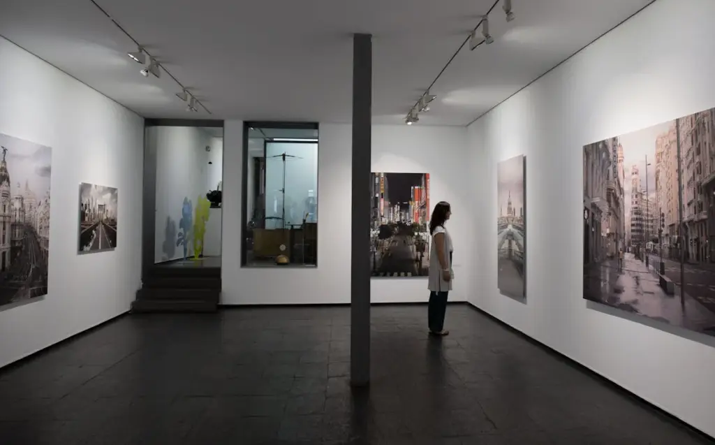 A person stands in an art gallery, observing large photographs of urban scenes. The room is well-lit with white walls and dark flooring.