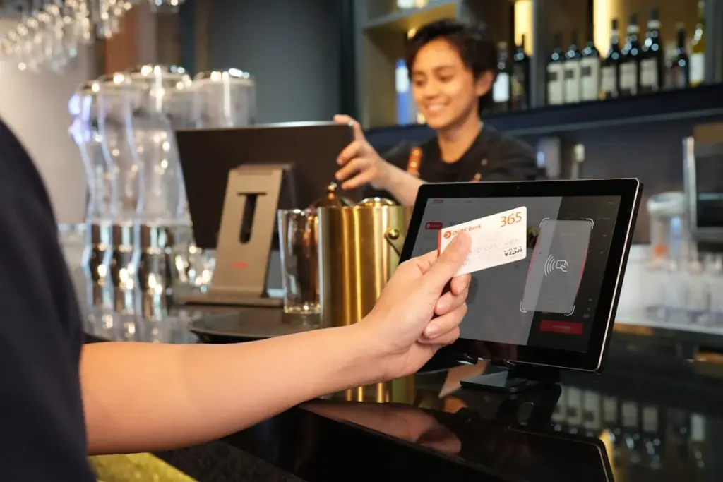 A person holds a card near a screen for a contactless payment at a bar, with the bartender in the background. Americans are tired of tipping, and this cashless option eases tipping fatigue for many customers.