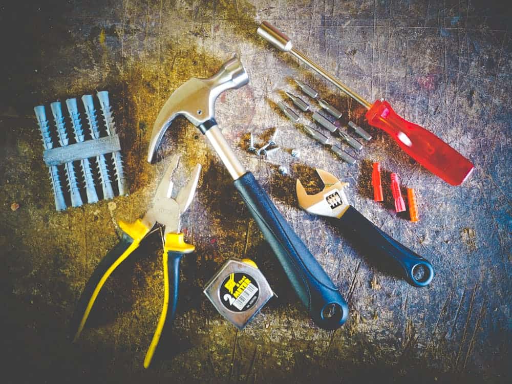 Various tools including a hammer, pliers, screwdriver, adjustable wrenches, a measuring tape, wall anchors, and drill bits are arranged on a worn surface—perfect examples of things you're paying for but can get for free if you know where to look.