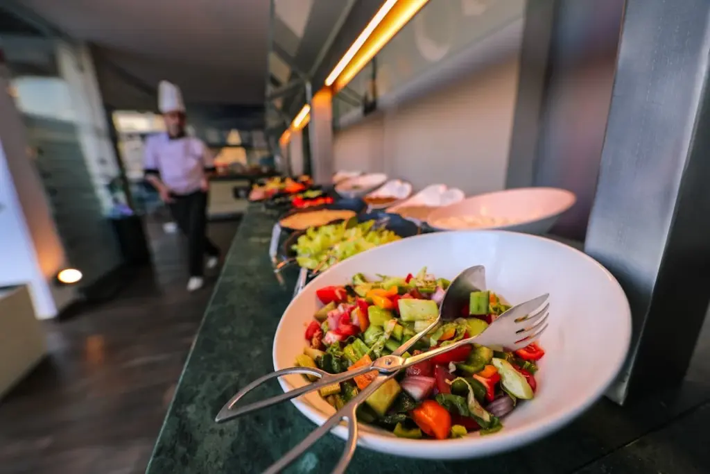 A salad bar with various fresh vegetables in a bowl in the foreground and a person wearing a chef's hat in the background, emphasizing that this is one of those places where it is not recommended to leave a tip.