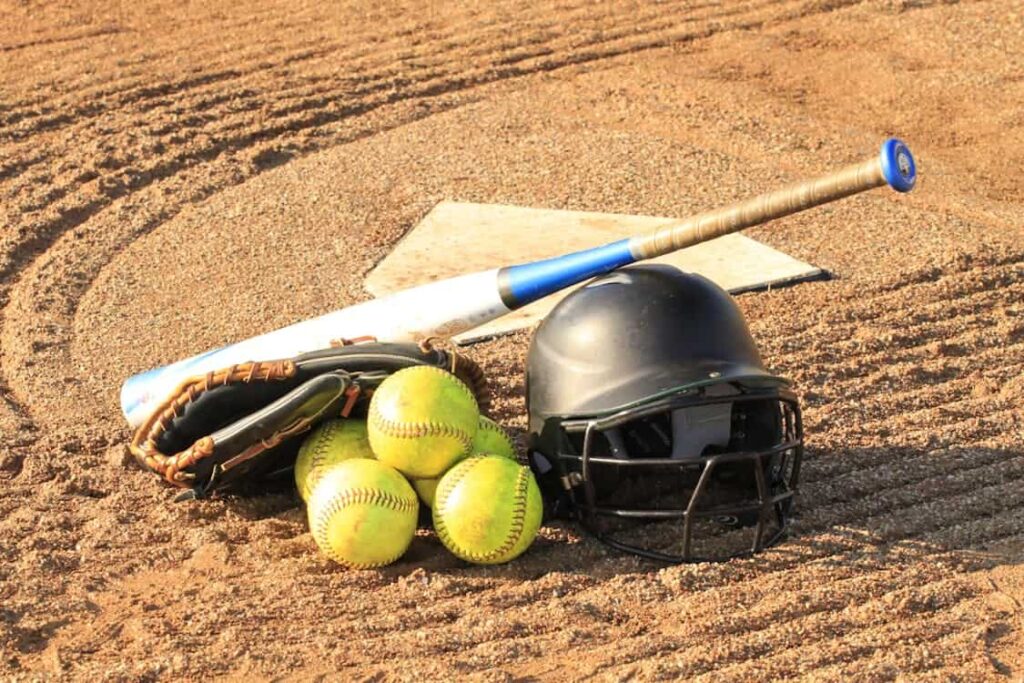 A softball bat, a glove, a helmet, and four softballs are scattered on a dirt field with home plate visible in the background, capturing the essence of lower-middle-class recreation.