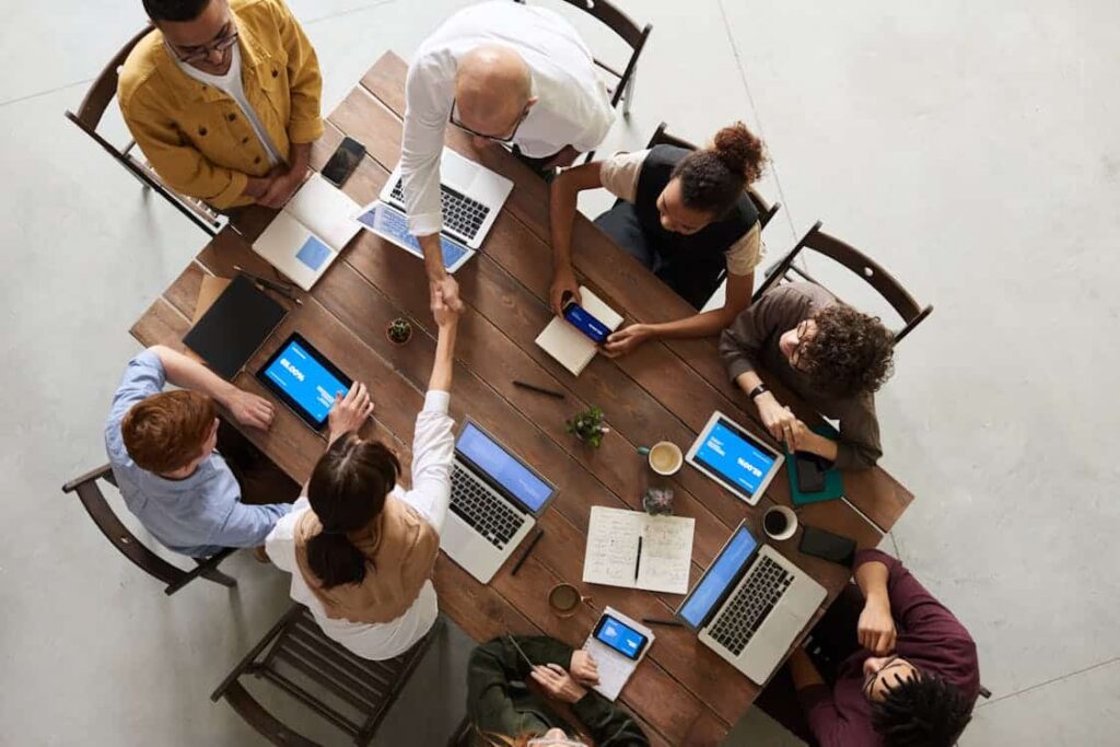 Aerial view of a diverse group of people sitting around a wooden table filled with laptops, notebooks, and phones, engaged in a meeting; two individuals, acknowleding the things replaced by technology, are shaking hands.
