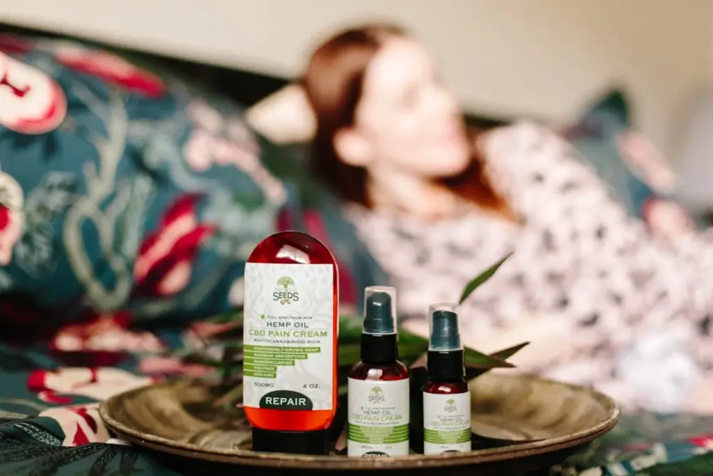 A blurred person lies on a bed in the background while a tray in the foreground holds bottles of hemp oil and CBD pain cream labeled "Repair".