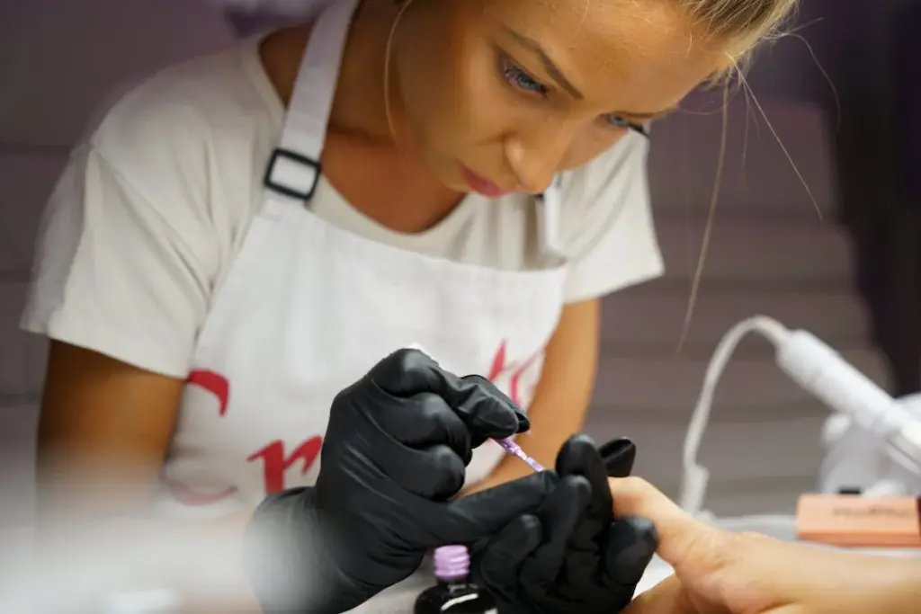 A woman in an apron and black gloves is carefully applying nail polish to another person's fingernails, one of those places where it is not recommended to leave a tip.
