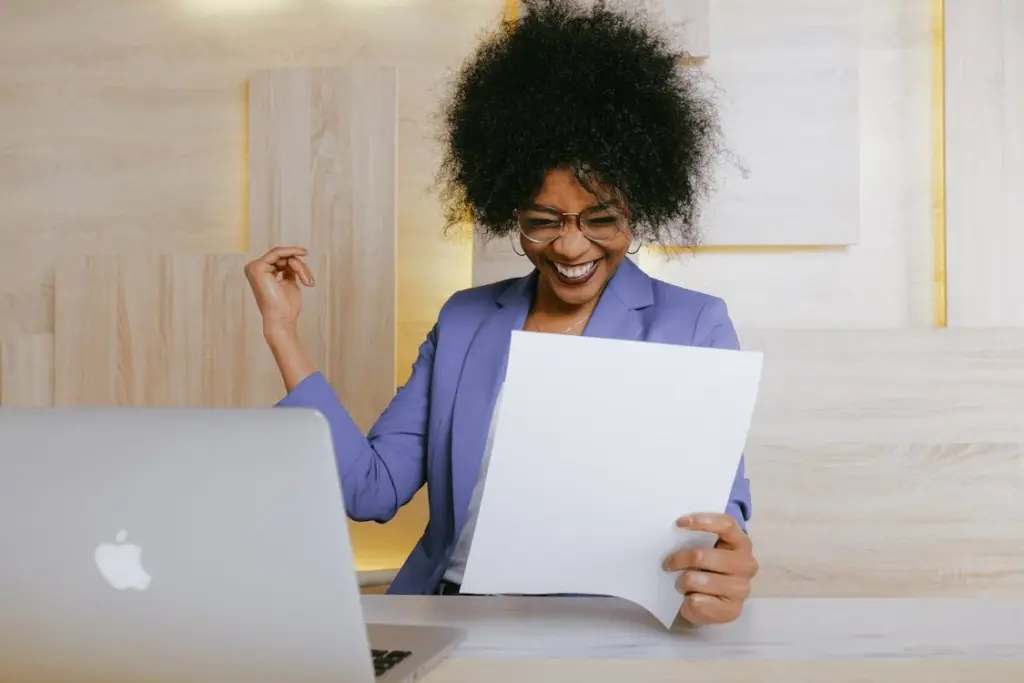 A person wearing glasses and a blue blazer, indicative of the lower middle class, smiles while reading a document at a desk with a laptop.