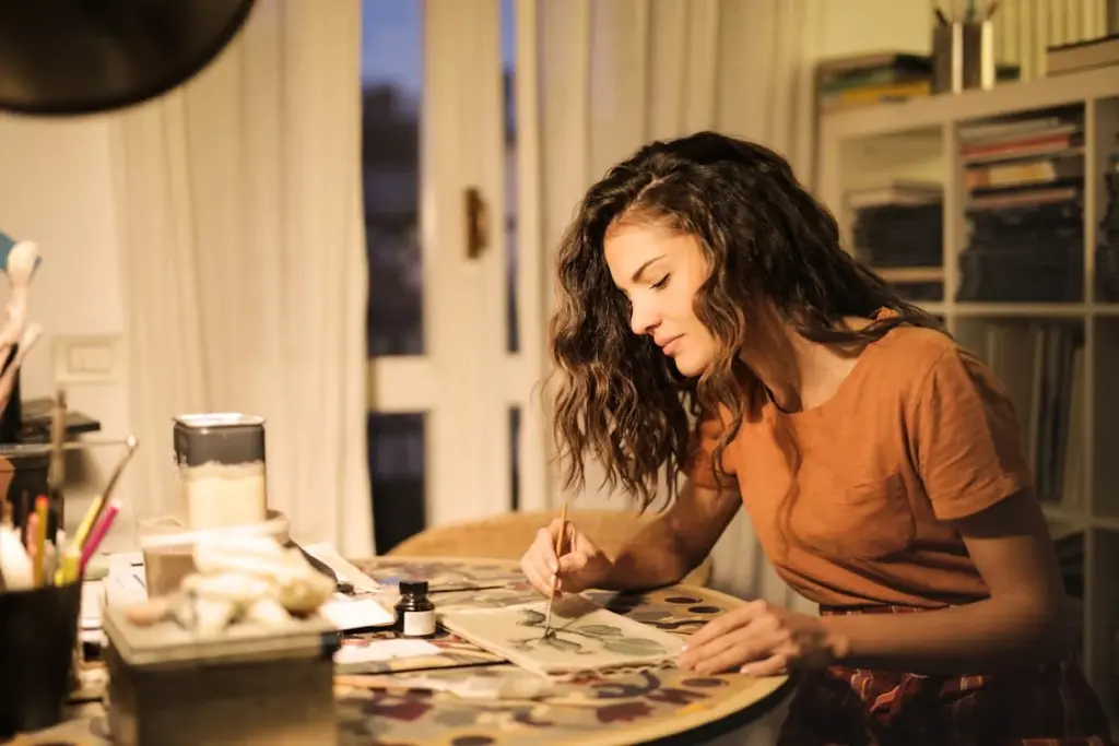 A woman with wavy hair and a brown shirt sits at a table, painting on a canvas with various art supplies around her.