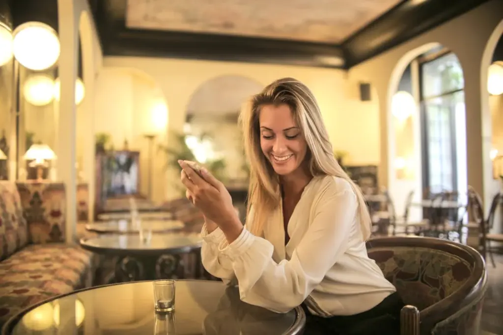 A woman with long blonde hair and a white blouse sits at a glass table in a warmly lit café, smiling while looking at her phone, the screen reflecting the myriad of things replaced by technology.