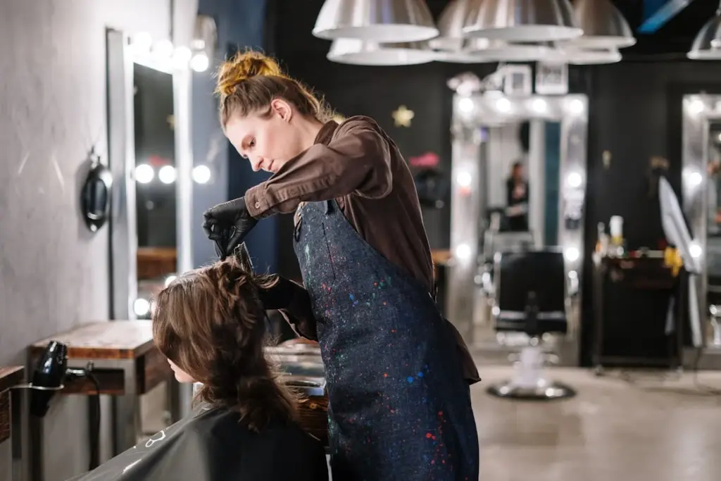 A hairdresser wearing an apron and gloves styles a client's hair in a modern salon with bright lighting, one of those places where it is not recommended to leave a tip.