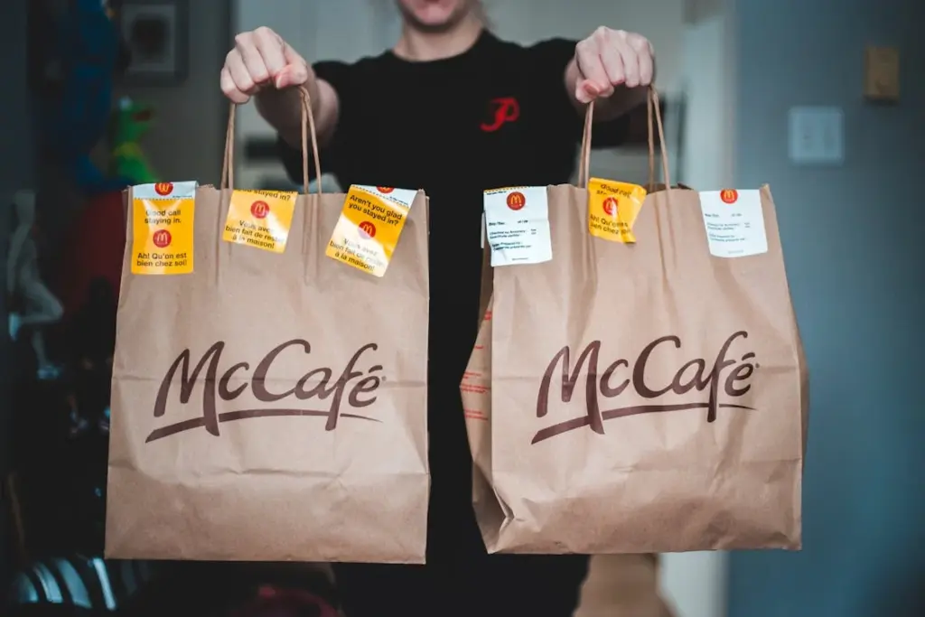 Person holding two McDonald's McCafé paper bags filled with food orders, each with receipts attached, in a home environment. Amid growing tip fatigue, many Americans are tired of tipping for takeout and delivery services.