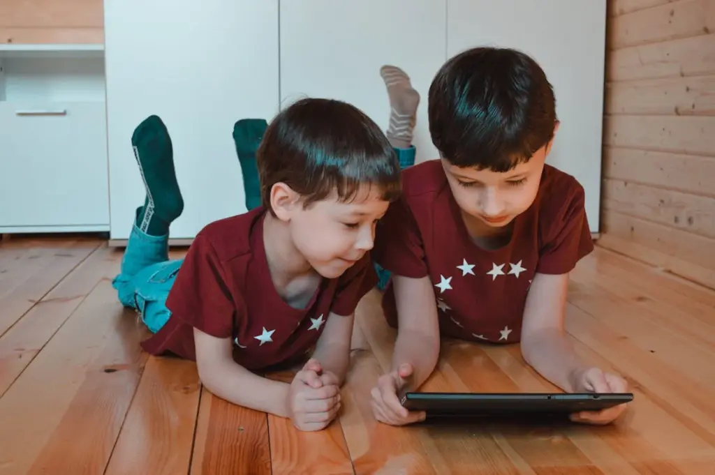 Two children, dressed in matching red star-patterned shirts, lie on a wooden floor in a wood-paneled room, engrossed in a tablet together—one of the many things replaced by technology.