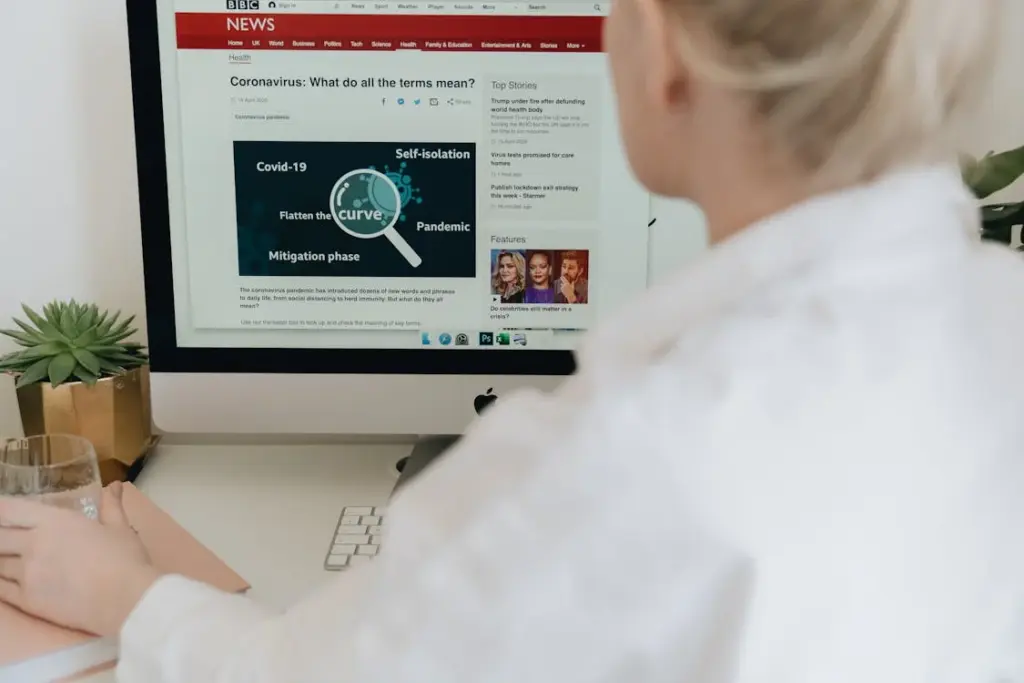 A person in a white shirt is reading a BBC News article about coronavirus terms on a desktop computer screen. A glass of water, a notebook, and a potted plant are on the desk—simple comforts resisting things replaced by technology.