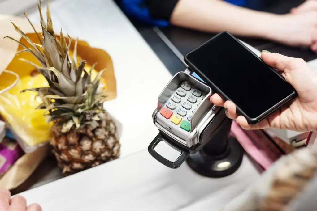A person holds a smartphone with a payment app open near a card reader at a checkout counter, next to a pineapple and other groceries, showcasing how things replaced by technology have transformed everyday tasks.