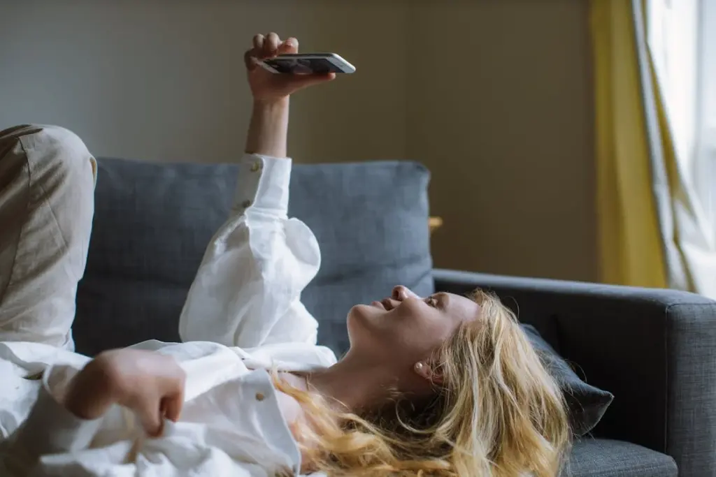 A person with long blonde hair in a white shirt lies on a gray sofa, holding a smartphone above their head and smiling, perhaps reflecting on all the things replaced by technology.